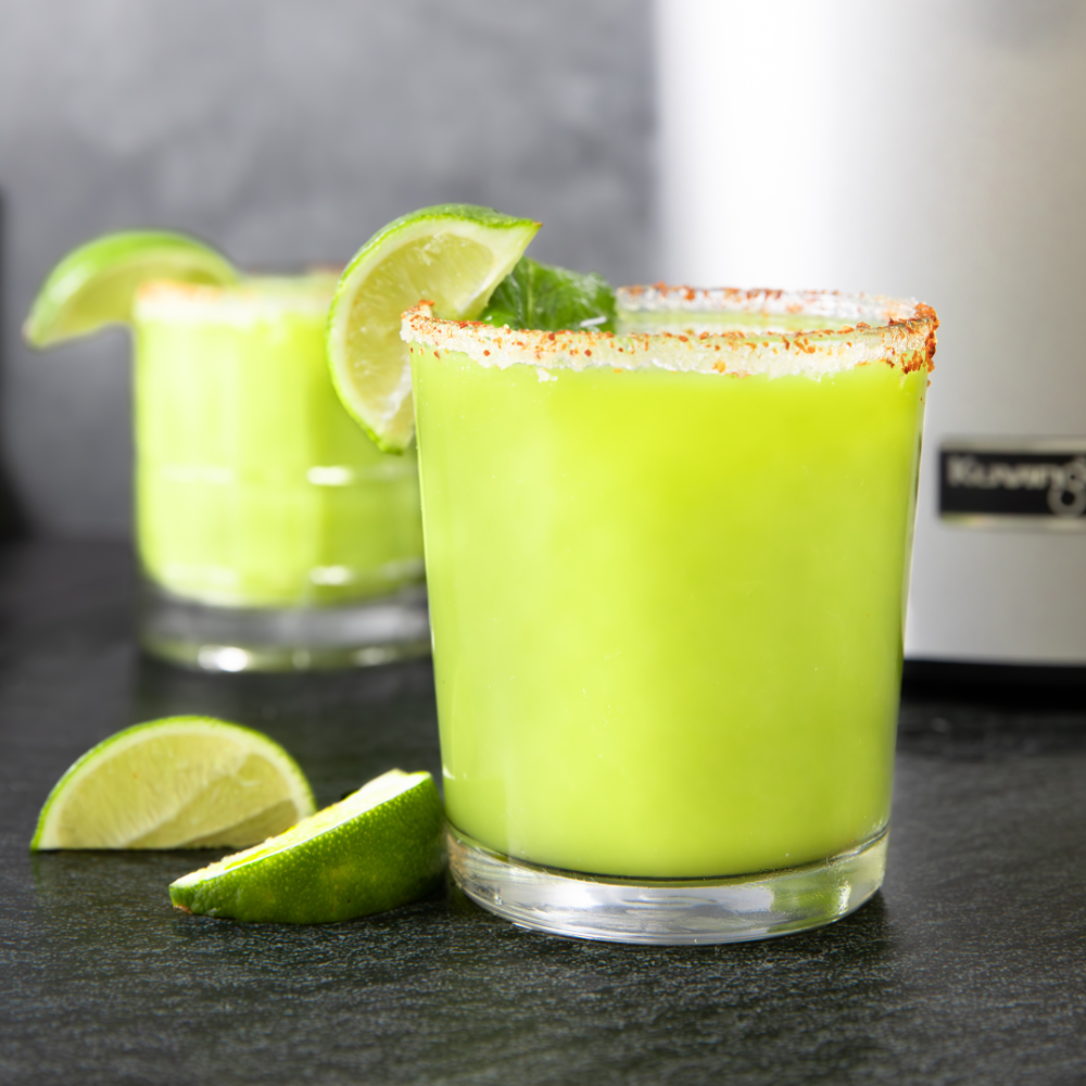 two glasses of green juice margarita and two slices of lime in front of a kuvings juicer